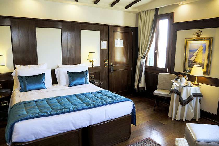 A hotel room with a queen bed featuring teal bedding, wood paneling, a small dining table, and framed nautical art.