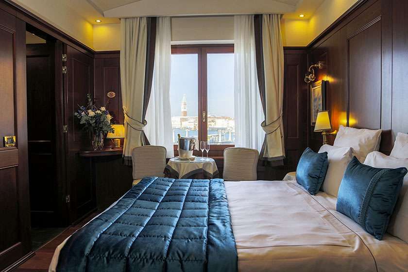 A luxury train cabin with a bed featuring blue bedding and teal pillows, and a window with a view of a city landscape.