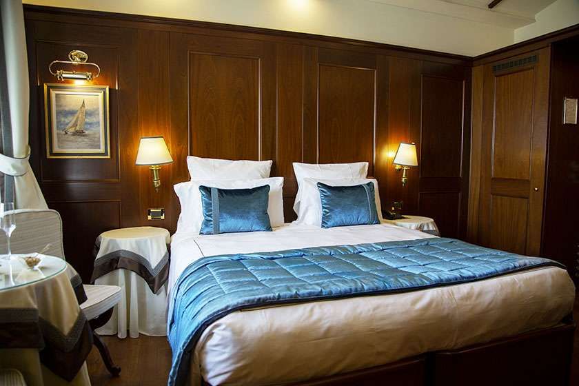 A hotel room featuring a queen bed with a blue quilted bedspread, dark wood paneling, and two wall-mounted lamps.