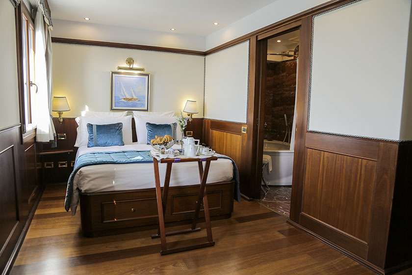 A cozy hotel room featuring a bed with blue accents, a wooden folding tray, and an adjacent bathroom with a tub.