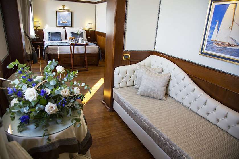 A luxury hotel suite featuring a bed, a tufted cream sofa, and a glass table with a large bouquet of white and blue flowers.