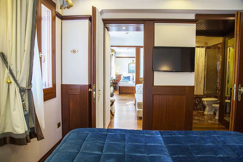 A view from a blue bed through a doorway into another room, with a wall-mounted television and an adjacent bathroom.