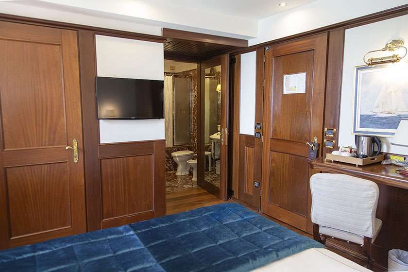 A hotel room with a blue-quilted bed, a wall-mounted TV, a wooden desk with a white chair, and an open bathroom door.