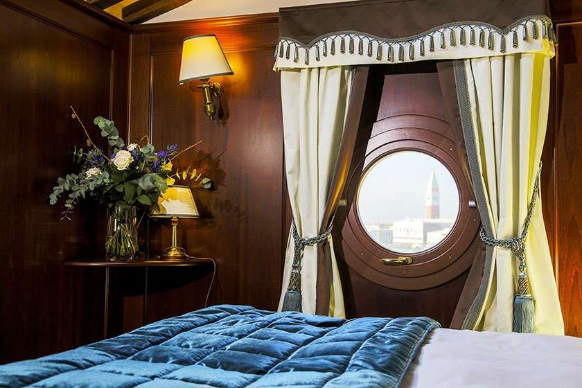 A blue bed in a wood-paneled room with a lamp, flowers, and a round window with draped curtains.