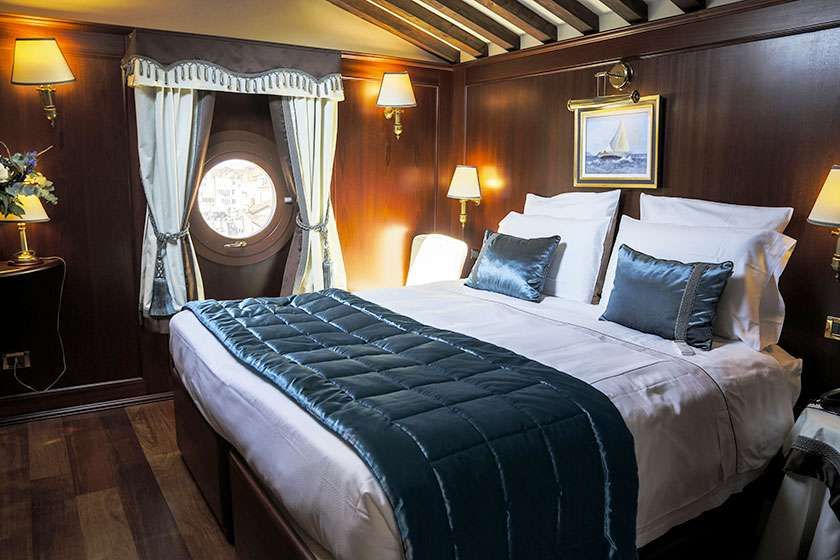 A cozy bedroom with dark wood paneling, a circular window with curtains, and a bed featuring a dark blue quilted runner.