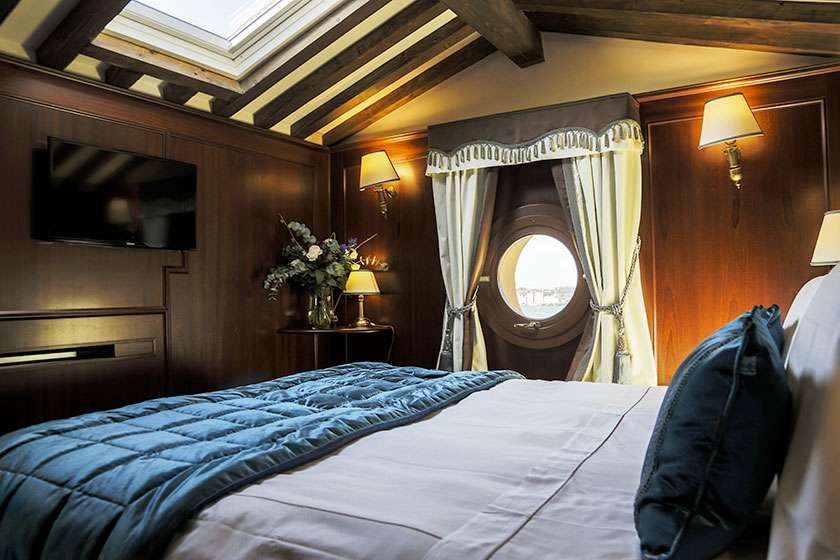 A luxurious cabin room featuring a bed with blue bedding, dark wood walls, a round window, and a skylight.