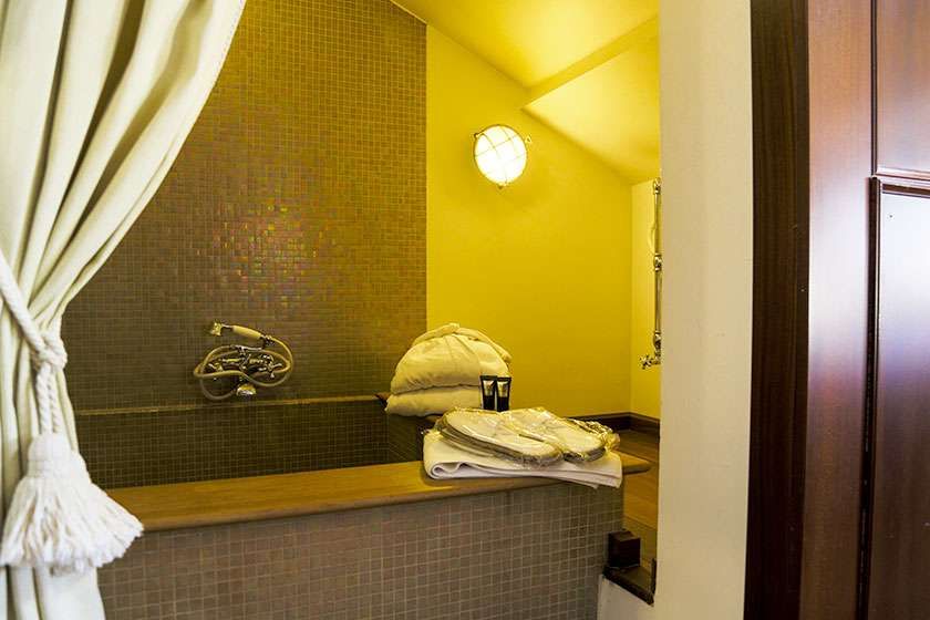 A bathroom with a tiled bathtub, a white curtain, folded towels, and slippers, lit by a circular wall lamp in yellow tones.