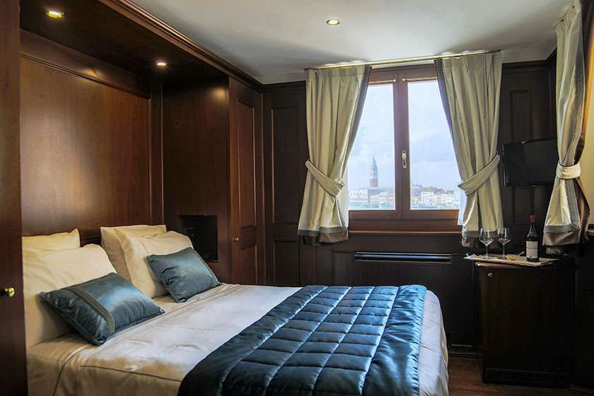 A cozy bedroom featuring a queen bed with white linens and a dark blue quilt, wooden walls, and a window overlooking a city.