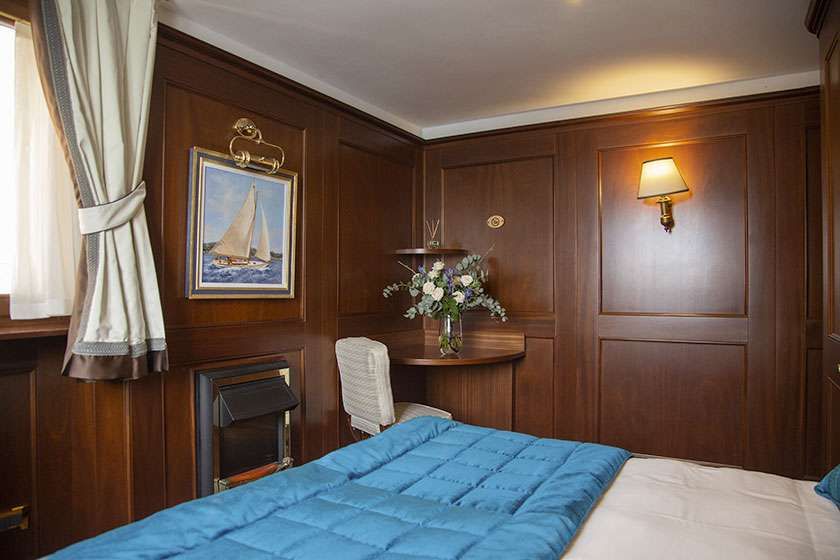 A wood-paneled cabin with a bed featuring a blue quilted comforter, a white chair at a small desk, and a sailboat painting.