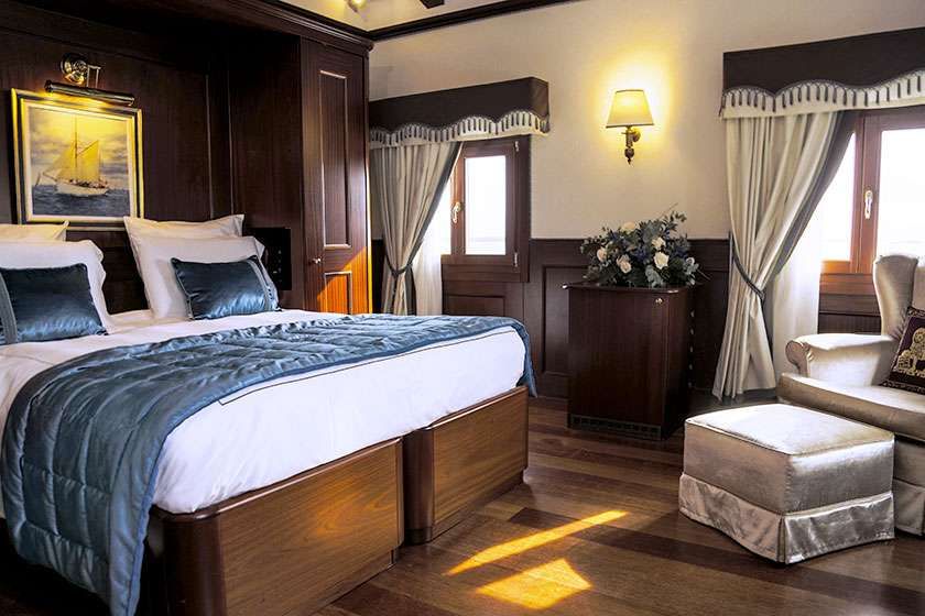 A luxurious bedroom with a large bed, blue bedding, wood accents, windows with white curtains, and a beige armchair.