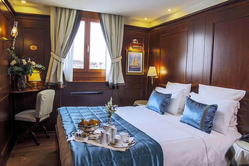 A luxury hotel bedroom with dark wood paneling, a bed with blue bedding and pillows, a breakfast tray, and a window.