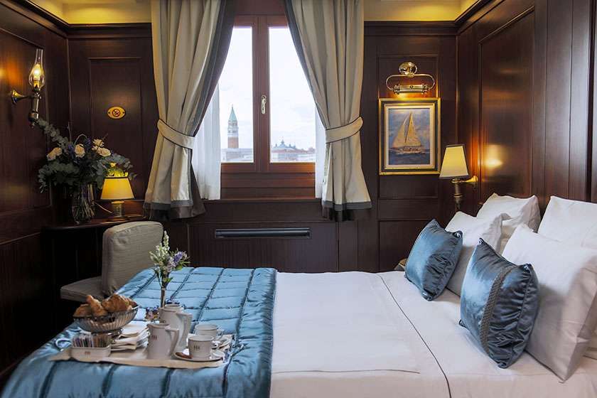 A cozy, wood-paneled hotel room with a queen bed, blue bedding, and a view of a Venetian bell tower through the window.