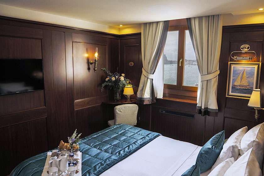 A cozy, wood-paneled hotel room with a queen bed, blue bedding, a desk, a window with curtains, and a sailboat painting.