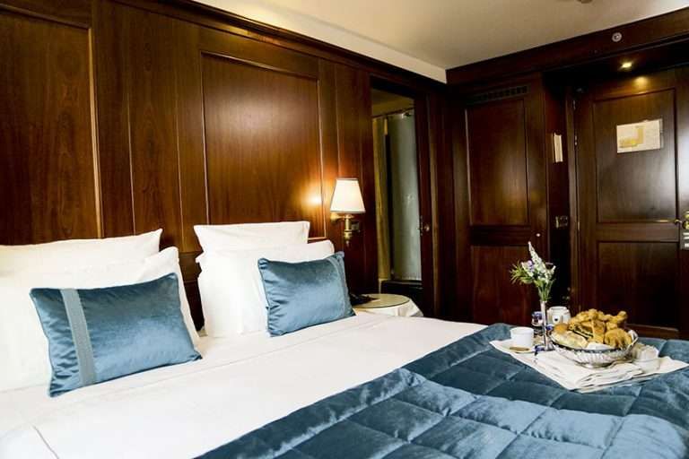 A luxury hotel room featuring a bed with white linens and teal accents, dark wood paneling, and a small fruit bowl on top.