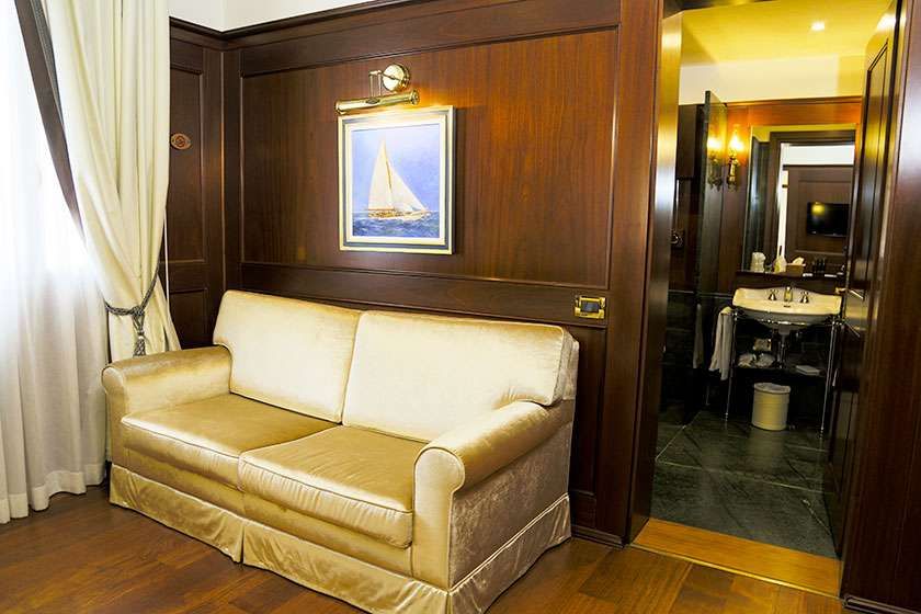 A cream-colored sofa sits in a wood-paneled room beneath a sailboat painting, with an open bathroom visible to the side.