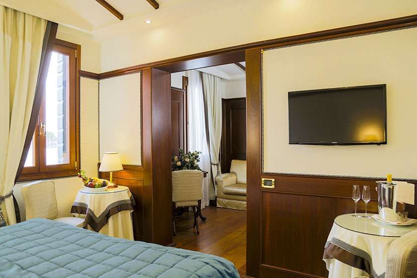 A hotel bedroom with a blue quilt, a flat-screen TV on the wall, and a side table with glasses and an ice bucket.