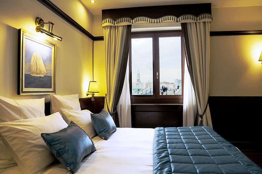 A hotel room with a queen bed featuring blue pillows and a quilted cover, next to a window with neutral-toned curtains.