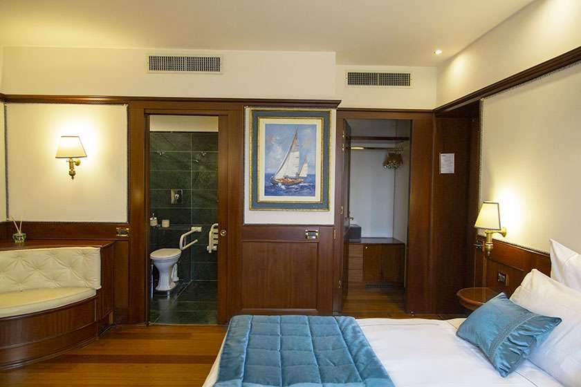 A bedroom with a bed, wood-paneled walls, a small lounge area, a bathroom door, and a closet, decorated with a boat painting.