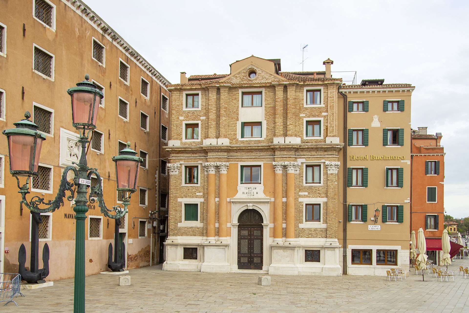 A view of the ornate San Biagio church facade in Venice, flanked by tall, sunlit historic buildings and a lamp post.
