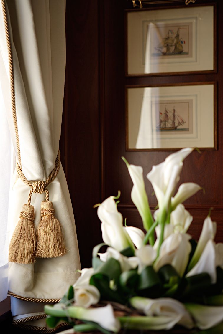 A lush bouquet of white calla lilies sits beside an off-white curtain with golden tassels and framed nautical art.