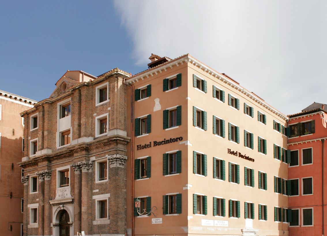 A tan-colored Hotel Bucintoro with green shutters stands beside a historic, weathered brick building in Venice.