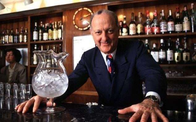 A person in a suit stands behind a bar, hands resting on the counter next to a large, ice-filled glass pitcher.