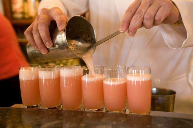 A bartender pours a frothy, pink cocktail from a metal shaker into a row of glasses lined up on a bar counter.