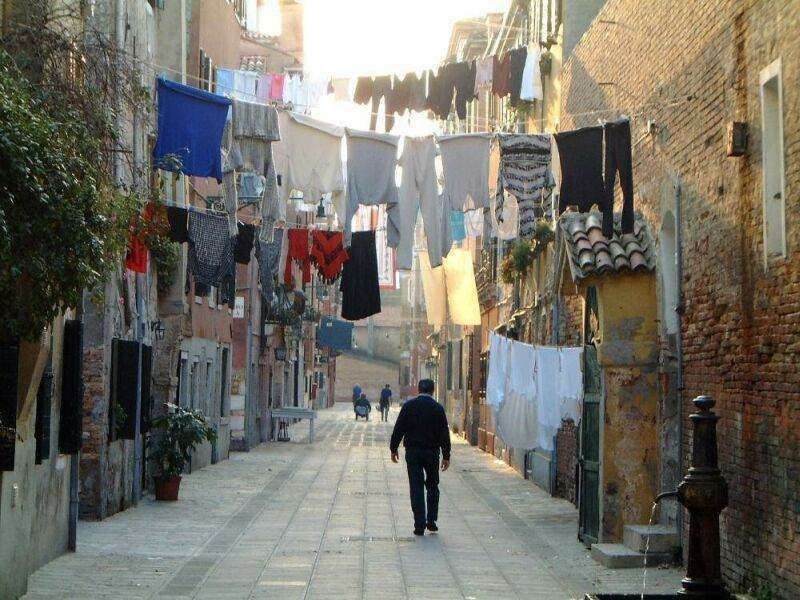 A person walks down a narrow, sunlit stone alley in Venice, with clothes hanging from lines overhead between brick walls.
