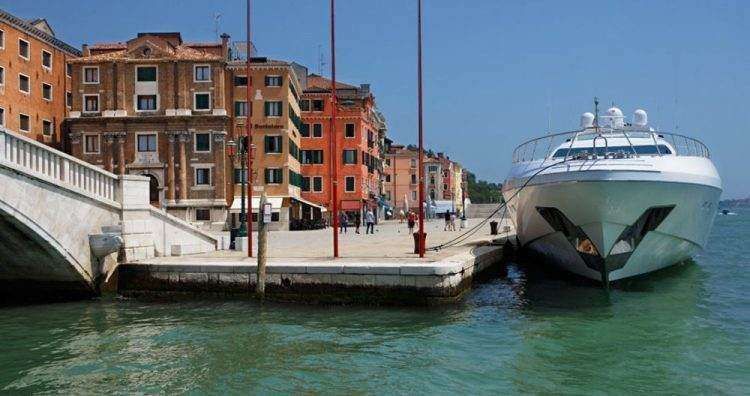 A large white yacht docked next to a stone quay in front of tall, orange-colored buildings on a sunny day.