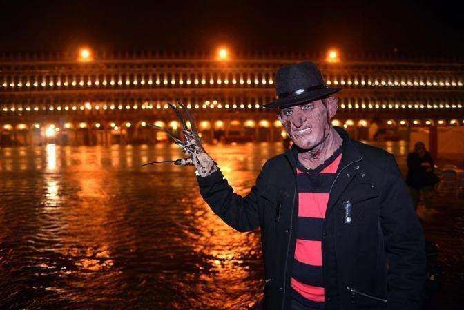 A person in a Freddy Krueger costume stands in a flooded square at night, holding up a clawed hand with the city behind.