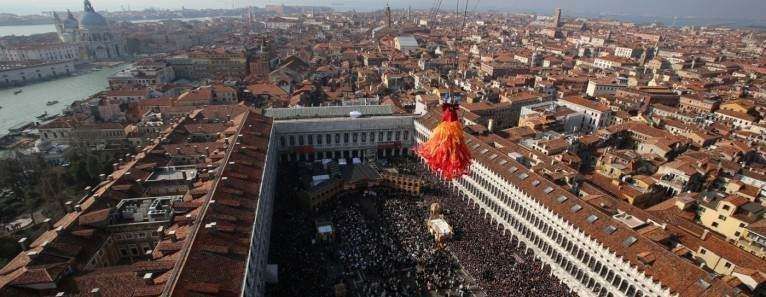 An aerial view of Venice as a performer in a large, vibrant red and orange dress descends toward St. Mark's Square.