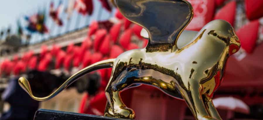 A golden statue of a winged lion, the Venice Film Festival award, set against a blurred red and white background.
