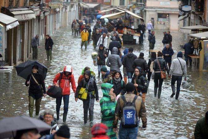 People navigate a flooded European street filled with water, walking past shop stalls and wearing casual clothing.