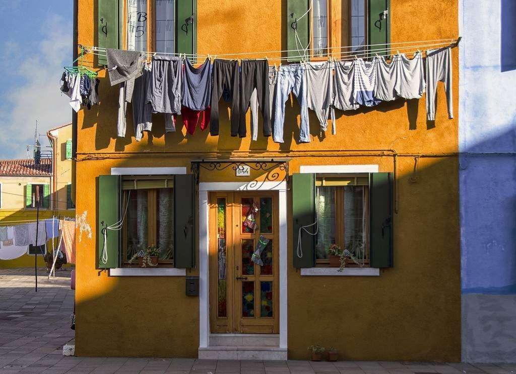 Laundry hangs to dry on a line across the facade of a vibrant yellow house with green shutters on Burano, Italy.