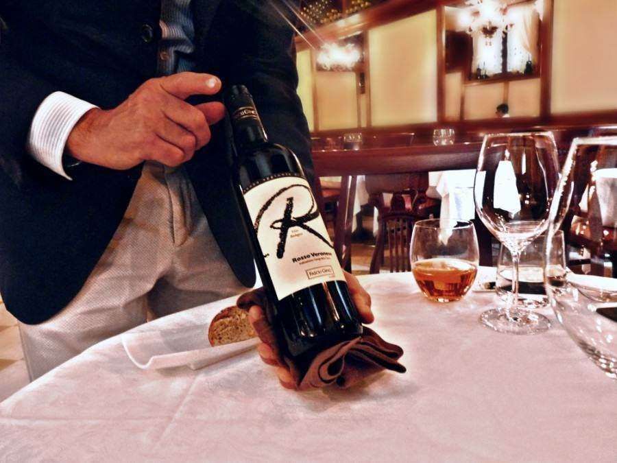 A server presents a bottle of red wine with a large letter R on the label at a restaurant table.