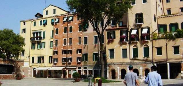 Two people walk through a sunny, paved Venetian square surrounded by historic multi-story buildings and a large tree.