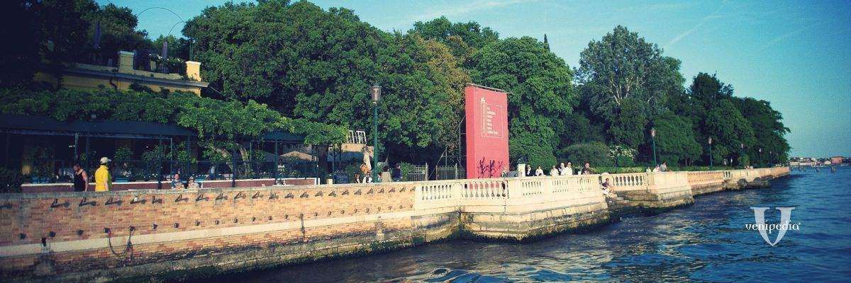 A scenic waterfront walkway bordered by trees and a stone balustrade, featuring a prominent red vertical sculpture.