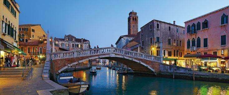 A stone footbridge arches over a narrow canal in Venice at dusk, flanked by historic buildings and soft golden lights.