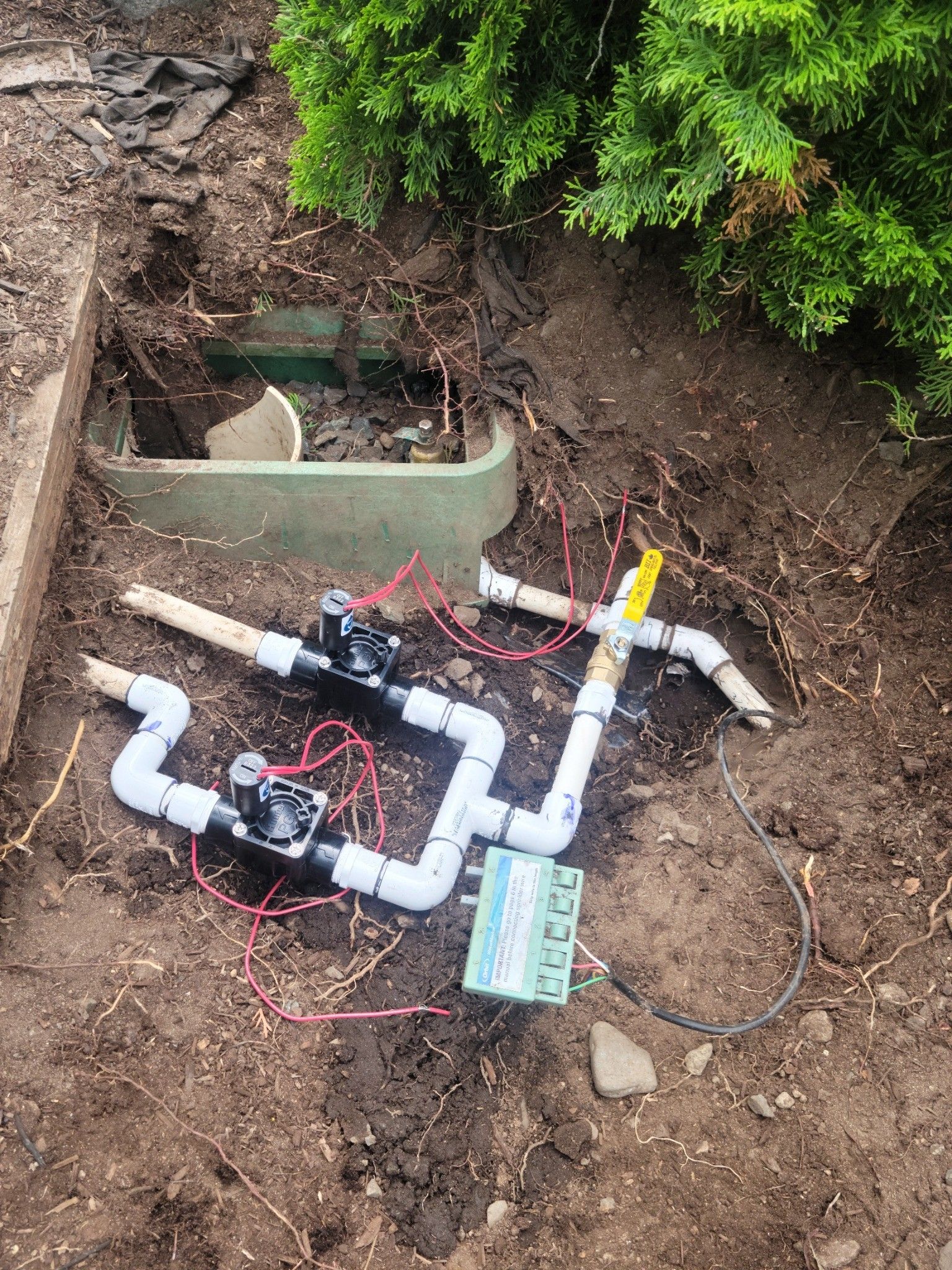 Exposed underground pipes and valves in a dirt trench beside a concrete box and greenery