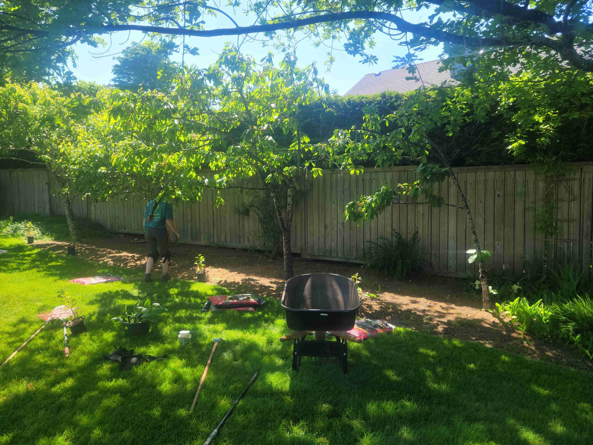 A man is standing in a backyard with a wheelbarrow and a grill.