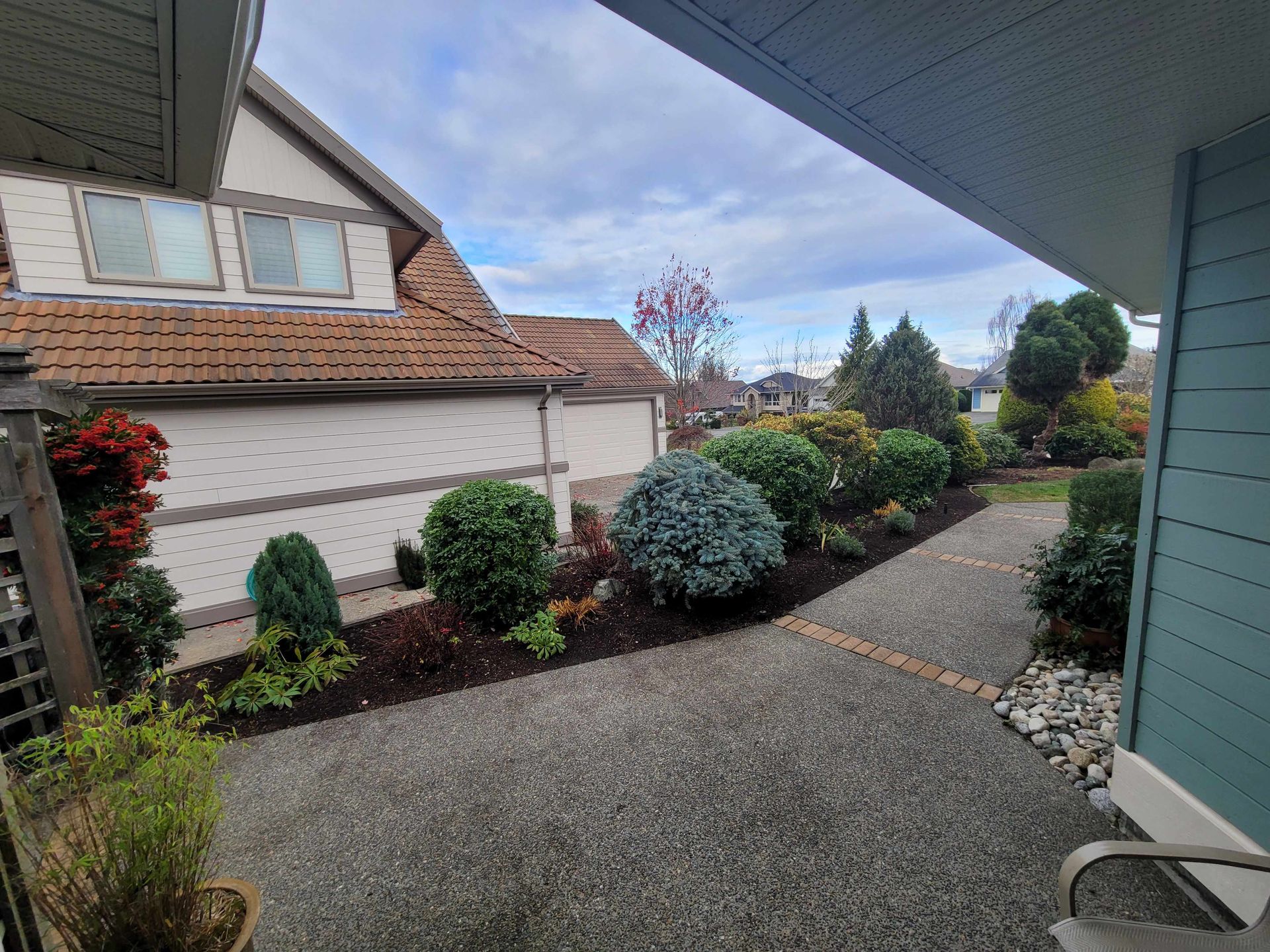 Front yard path beside a house with shrubs, flowers, and a brick garage under a cloudy sky