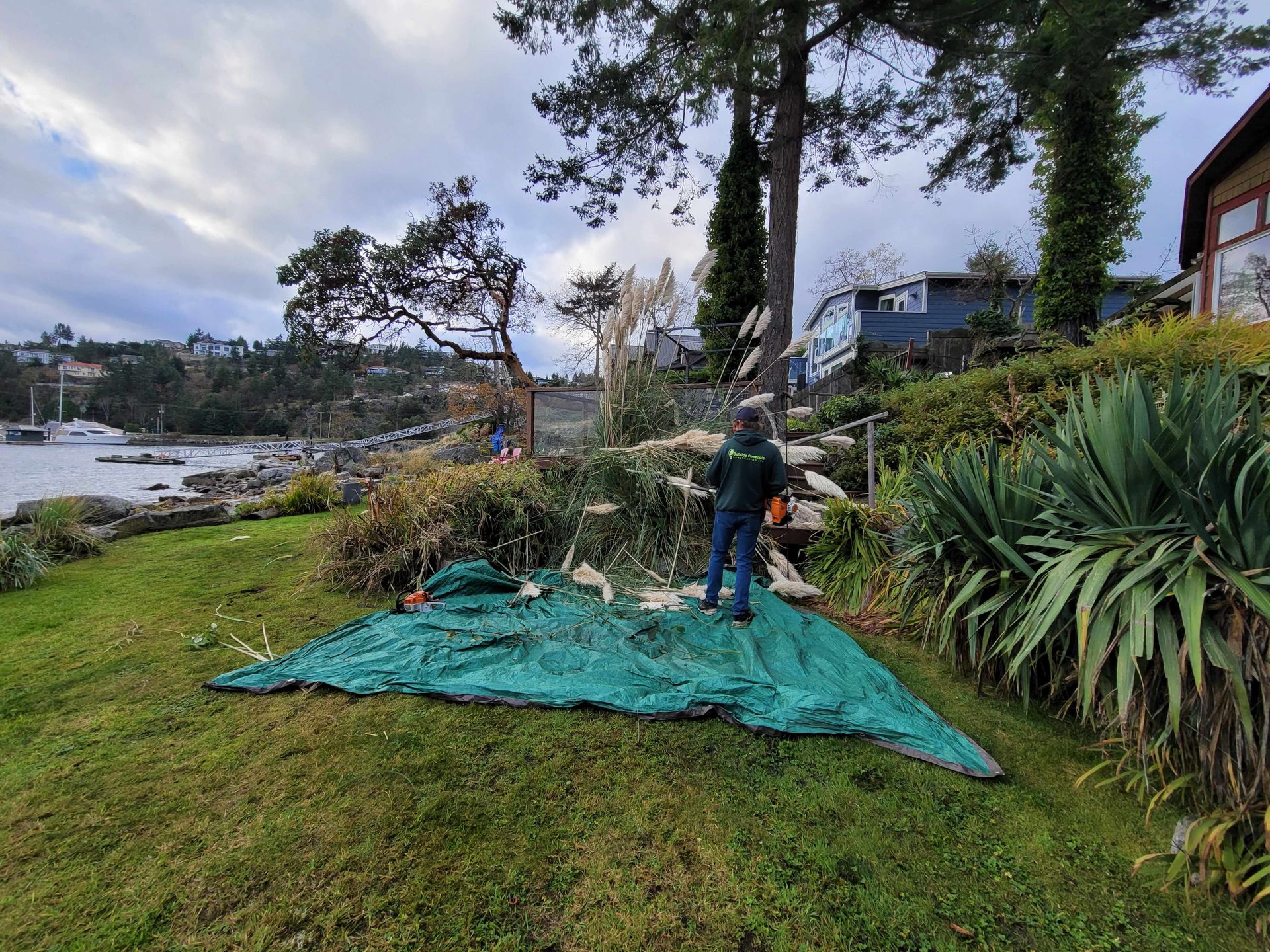 Person setting up green tarp on grassy riverbank near shrubs and trees