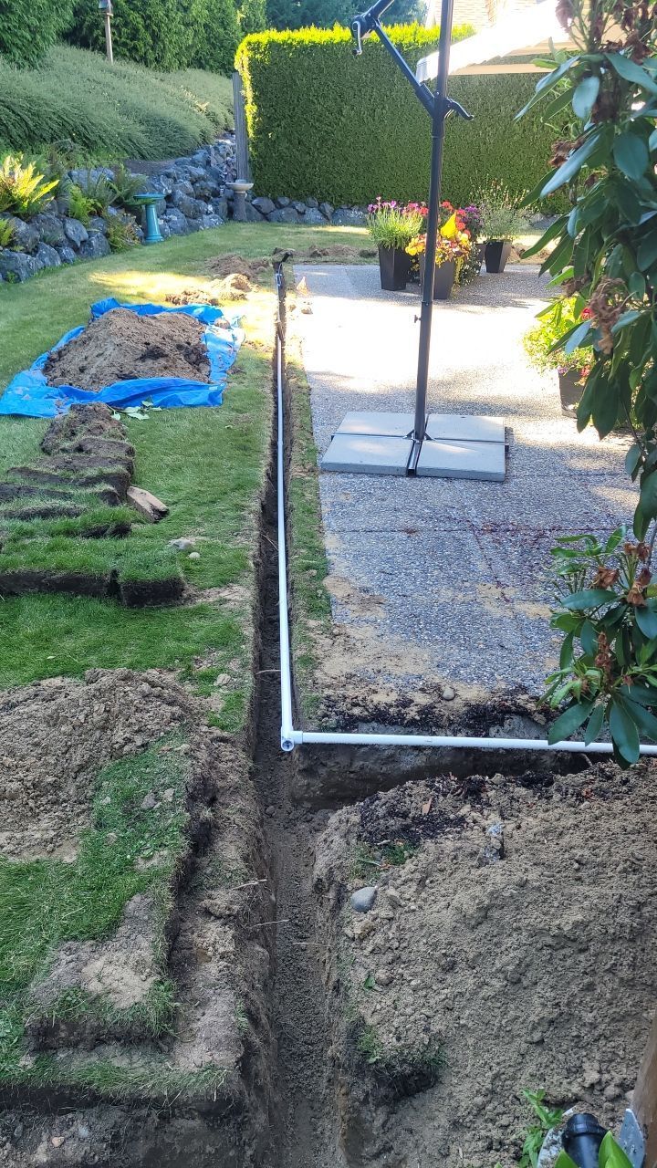 A trench dug in a backyard, with a white pipe running through it. Grass and soil are visible.