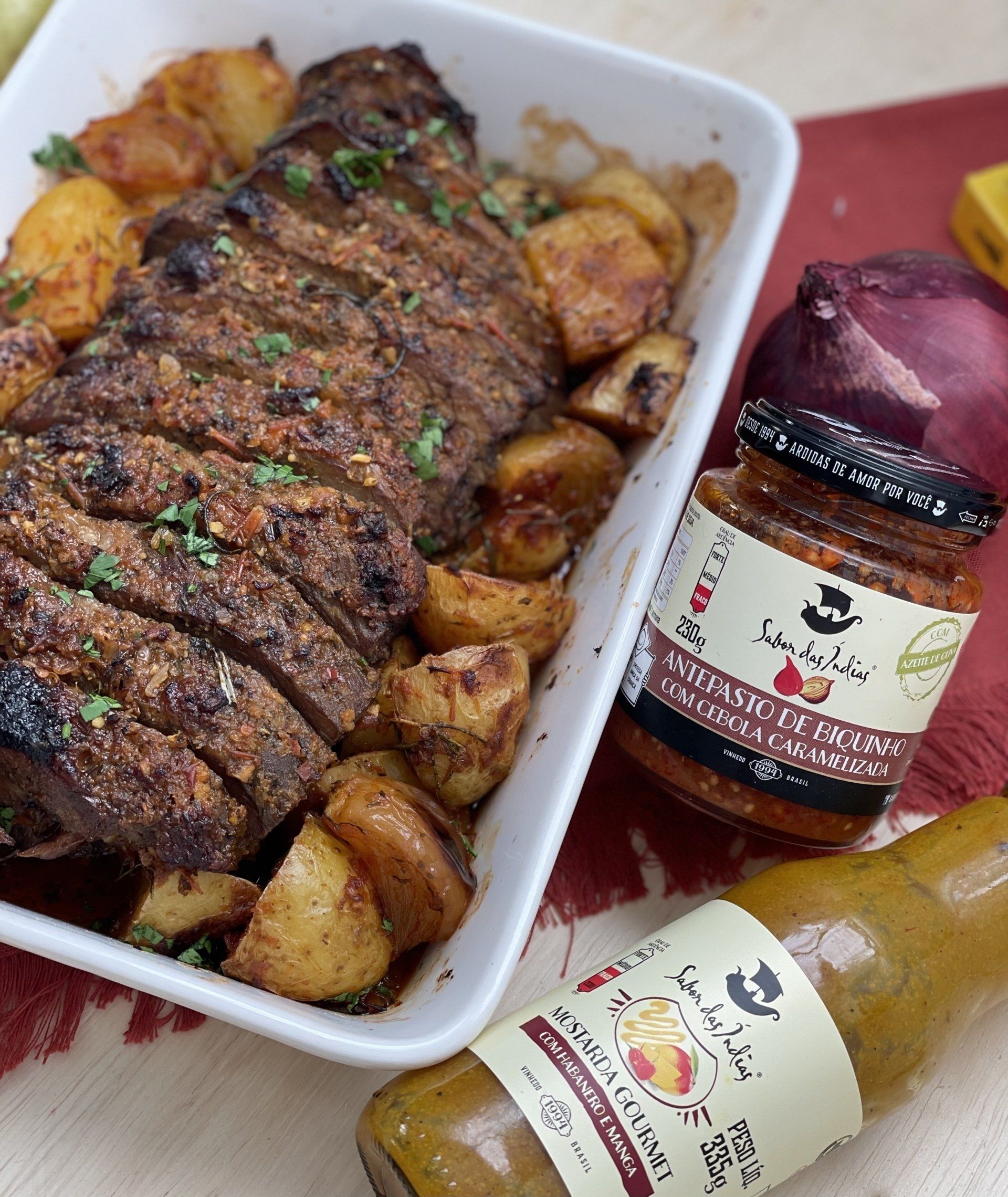 Carne assada em recipiente branco com batatas douradas na borda. A direita dois molhos.