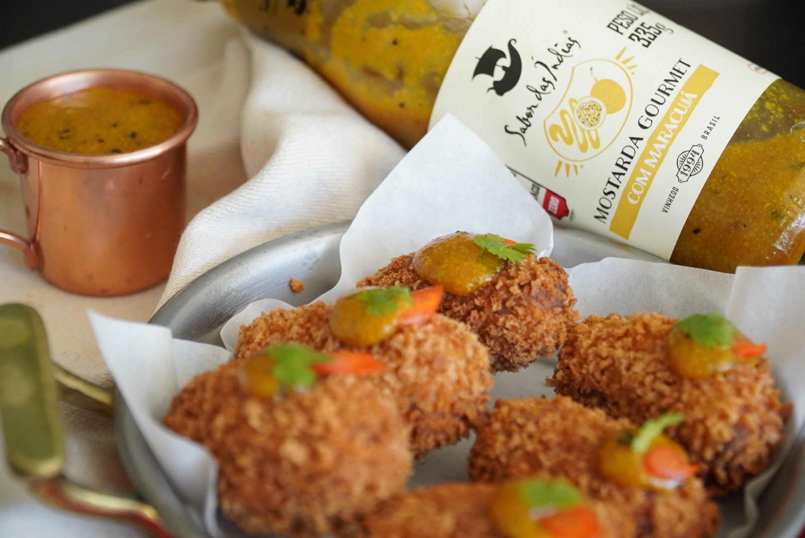 Da direita para esquerda, de trás para frente. Mostarda gourmet com maracujá, na diagonal. Bolinhos de carne (croquete) sob guardanapos brancos. Um pouco de cheiro verde e cenoura em cima.