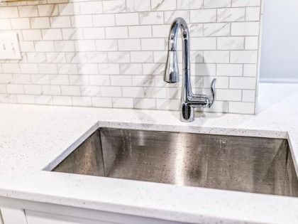 Stainless steel kitchen sink with chrome faucet against white countertop and tiled backsplash.