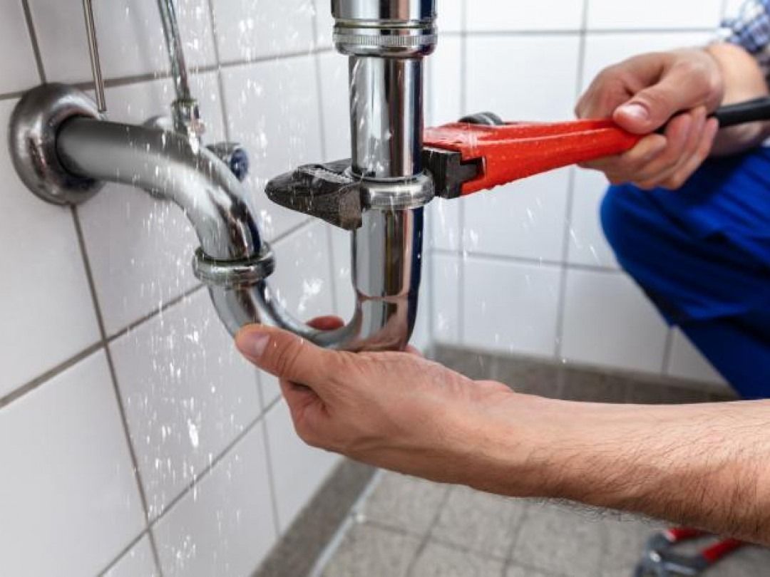 Plumber fixing a leaky sink pipe with a wrench; water is spraying.