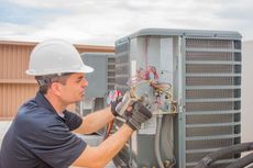HVAC technician in hard hat repairing outdoor air conditioner unit, using tools on a rooftop.