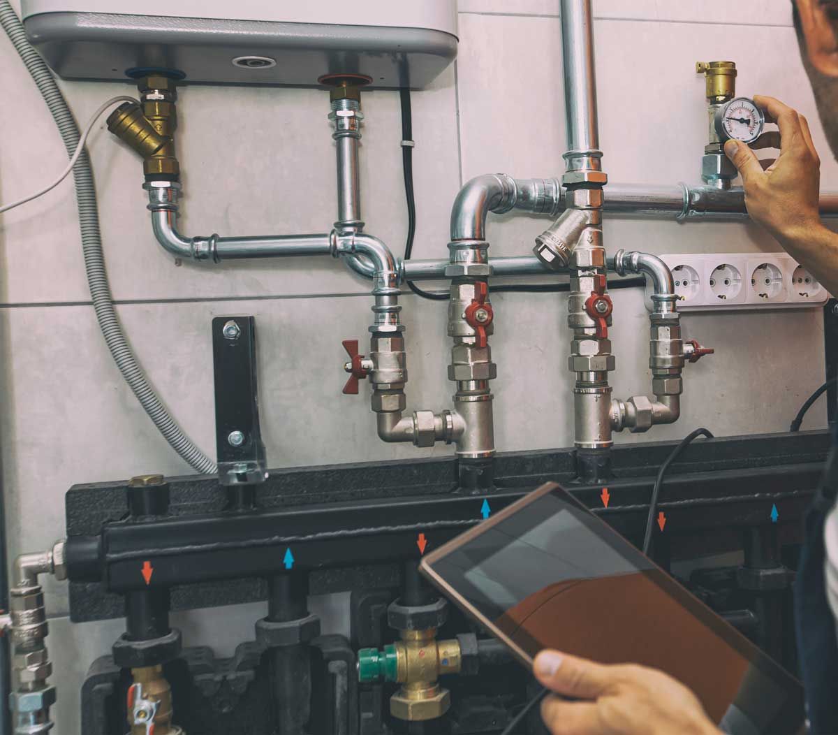 Plumber checking pressure gauge with a tablet, examining pipes near a boiler.