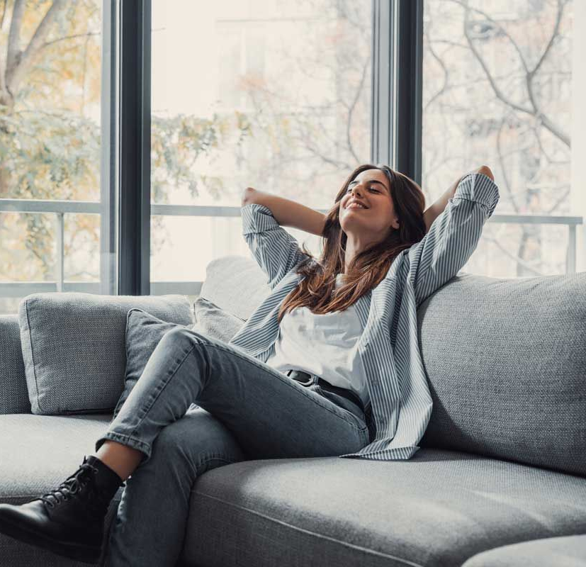 Woman smiling, relaxing on a gray couch in front of a window, arms behind her head.